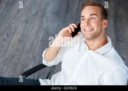 Sorridente imprenditore giovane seduto e parlando al telefono cellulare in ufficio Foto Stock