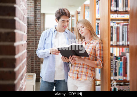 Giovani sorridente oriental l uomo e la giovane donna occidentale la condivisione di un libro nella biblioteca guardando verso il basso Foto Stock