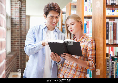 Giovani sorridente oriental l uomo e la giovane donna occidentale la condivisione di un libro nella biblioteca guardando verso il basso Foto Stock