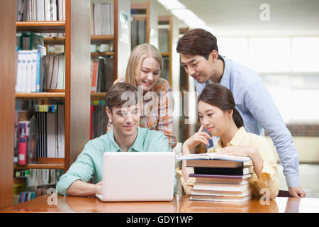 Quattro sorridendo gli studenti universitari che studiano insieme utilizzando un computer portatile e libri impilati in libreria Foto Stock