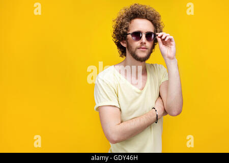 Ritratto di curly-pelose giovane uomo in occhiali da sole e bermuda cercando confuso a telecamera. Isolare. Sfondo giallo. Foto Stock
