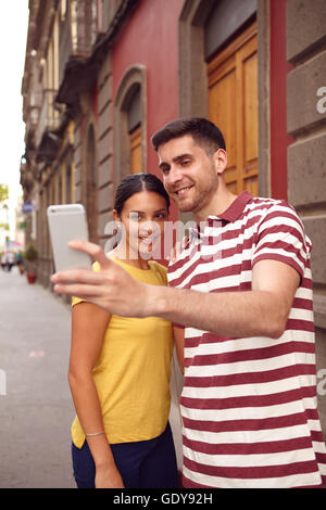 Coppia giovane guardando il telefono cellulare mentre sorridente gioiosamente per prendere un selfie, vestito casualmente in t-shirt con vecchi edifici behi Foto Stock