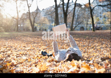 Donna matura libro lettura sdraiato sulla caduta foglie nel parco, Baviera, Germania Foto Stock