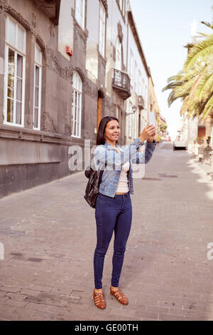 Piuttosto casualmente vestito giovane ragazza di scattare una foto con il suo telefono cellulare in una stretta strada pavimentata con edifici grigi dietro Foto Stock