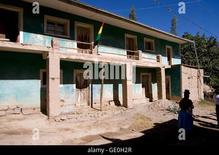 Bandiera boliviana di fronte ad un luminoso edificio rurale su una sterrata e una donna in pollera tradizionale nelle ombre Foto Stock