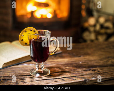 Vin brulé e un libro sul tavolo di legno. Camino con fuoco caldo sullo sfondo. Foto Stock