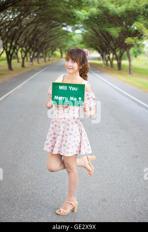 Carino donna mano azienda scheda verde segno con il testo ' ti sposerà me' sulla strada e albero, sorridente modello femminile. Foto Stock