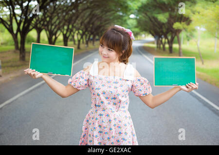 Carino donna mano azienda green blank board segno sulla strada con tree, sorridente modello femminile. Foto Stock