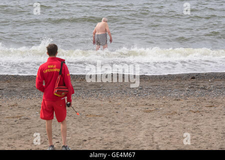 Un bagnino RNLI permanente sulla Aberystwyth Beach mantenendo un occhio guarda su un senior maschio adulto uomo nuotare nel mare, Regno Unito Foto Stock