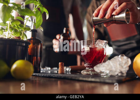 Immagine ravvicinata di barman versa manualmente da bere una tazza di misura in un cocktail di vetro riempita con cubetti di ghiaccio. Barista maschio preparata Foto Stock