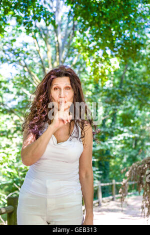 Curva elegante donna matura mette il dito indice alle labbra per chiedere silenzio in un giardino Foto Stock