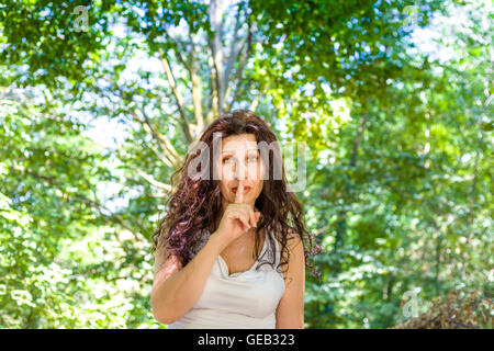 Curva elegante donna matura mette il dito indice alle labbra per chiedere silenzio in un giardino Foto Stock