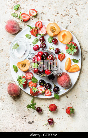 Una sana frutta estiva varietà. Ciliegie dolci, fragole, pesche, albicocche e foglie di menta in ceramica bianca che serve la piastra al di sopra della luce di sfondo in calcestruzzo Foto Stock