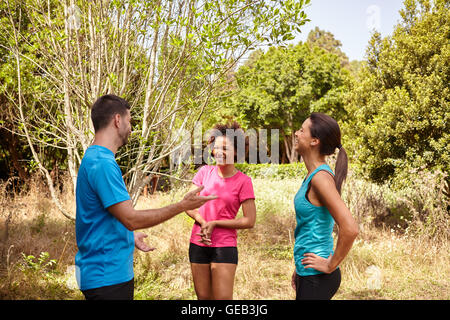 Un gruppo di tre giovani praticanti di jogging prendendo una pausa nella luce diurna e circondato da alberi e cespugli nella tarda mattinata di indossare la t-shirt e Foto Stock