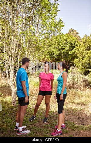 Tre giovani praticanti di jogging prendendo una pausa nella luce diurna e circondato da alberi e cespugli nella tarda mattinata di indossare la t-shirt e pan nero Foto Stock