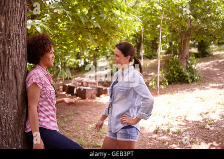 Due amiche ridendo e chiacchierando in la pezzata pomeriggio di sole mentre uno si appoggia contro un albero di indossare un abbigliamento informale Foto Stock