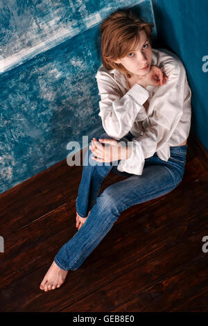 Lonely donna casual in camicia bianca e jeans blu seduta sul pavimento in un angolo di blu camera abbandonata. Vista superiore Foto Stock