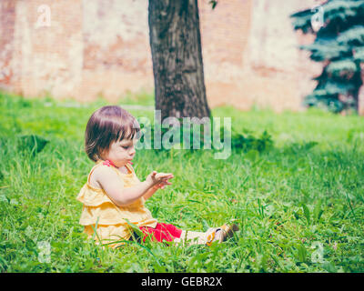 Poco ragazza seduta su erba guardando la sua mano Foto Stock