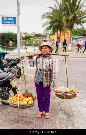 Hoi An, Vietnam - Febbraio 16, 2016: Asian venditore che trasportano la frutta fresca in ciotole sulle sue spalle in strada a Hoi An, Vietnam. Rambutan, mango, banana e mandarino. Foto Stock