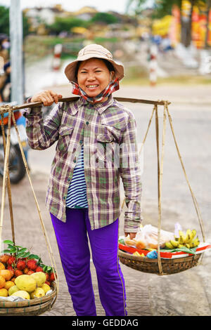 Hoi An, Vietnam - Febbraio 16, 2016: donna asiatica venditore che trasportano la frutta fresca in ciotole sulle sue spalle in strada a Hoi An, Vietnam. Rambutan, mango, banana e mandarino. Foto Stock