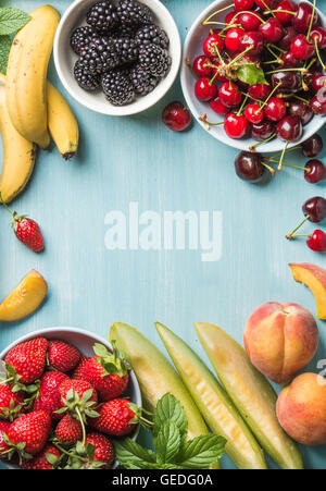 Una sana frutta estiva varietà. Ciliegie dolci, fragole, more, pesche, banane, fette di melone e foglie di menta su sfondo blu con una copia dello spazio nel centro, vista dall'alto Foto Stock
