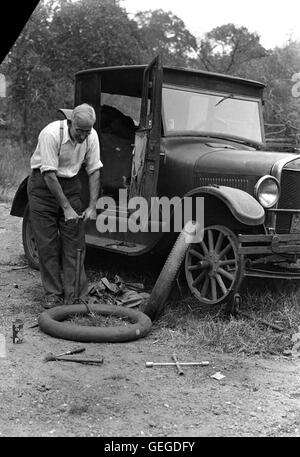 Viene mostrato un uomo che cambia uno pneumatico su un veicolo, dimostrando un compito comune del XX secolo. Questa pratica fotografia cattura un momento della vita quotidiana nei primi anni '1900 Foto Stock