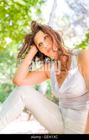 Bella pensieroso giovane donna indossa top bianco e pantaloni guardando la telecamera e tiene il capo in un stile di bellezza posano con le ginocchia fino in un parco Foto Stock