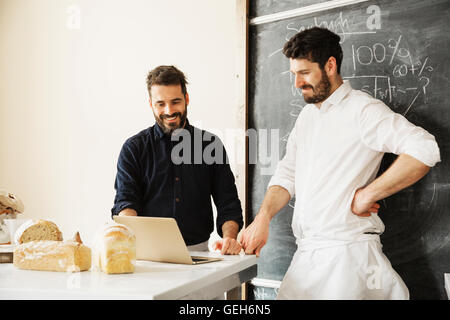 Due fornai sta in piedi in una tabella usando un computer portatile, pane appena sfornato, una lavagna a parete. Foto Stock