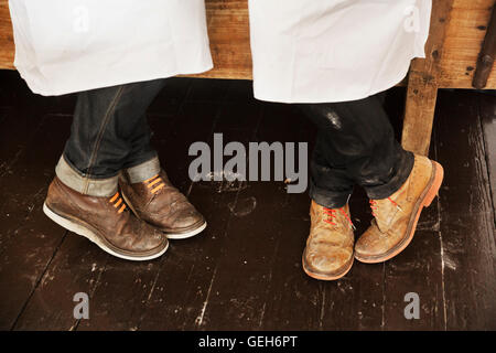 Chiudere fino a due uomini per le gambe e i piedi indossando grembiuli bianchi, jeans e brogues marrone rosso con lacci delle scarpe. Foto Stock