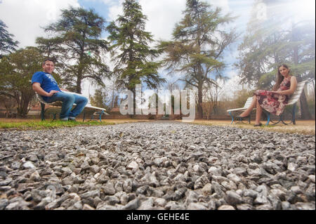 Bassa vista prospettica di un paio di seduta oltre sui banchi del parco con alberi di pino in background Foto Stock