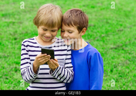 I bambini con il cellulare. Due ragazzi sorridenti, cercando di schermo, la riproduzione di giochi o utilizzando l'applicazione. All'esterno. Tecnologia istruzione tempo libero concetto di persone Foto Stock