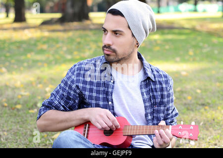 Ritratto di giovane uomo bello suonare il ukelele in un parco. All'esterno. Foto Stock