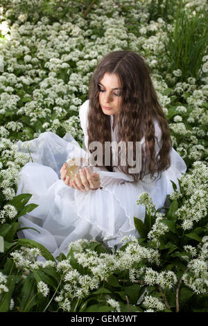 Giovane e bella donna che indossa un abito bianco in una foresta guardando nella sfera di cristallo Foto Stock