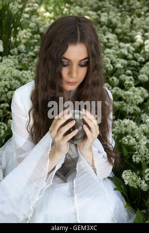 Giovane e bella donna che indossa un abito bianco in una foresta guardando nella sfera di cristallo Foto Stock