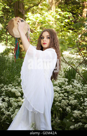 Bella donna che indossa un lungo abito bianco in piedi in una foresta tenendo un tamburello Foto Stock