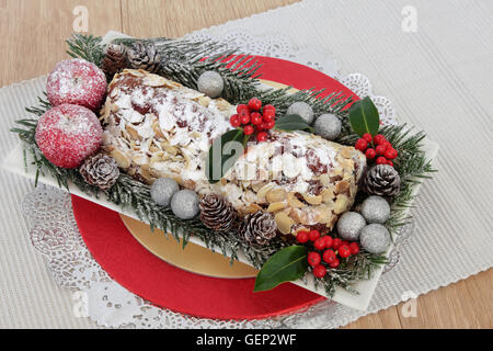 Lo Stollen la torta di Natale con holly, fir, decorativo mele e baubles di argento su un tavolo runner su oak. Foto Stock