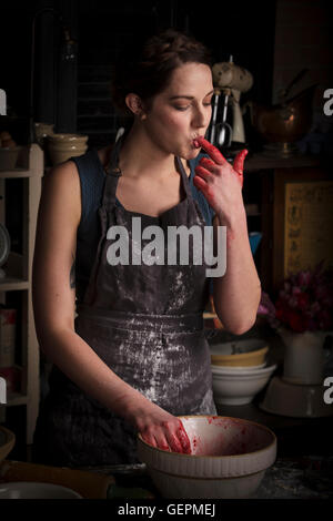 Il giorno di San Valentino la cottura, giovane donna in piedi in una cucina, la preparazione di confettura di lamponi, leccare le sue dita, degustazione. Foto Stock