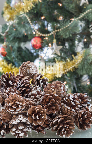 Verticale di close-up di pigne chiazzata con neve e un fuori fuoco, albero di Natale decorato in background Foto Stock