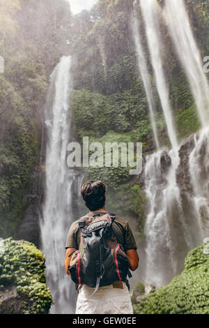 Vista posteriore del giovane uomo con zaino in piedi di fronte a cascata. Escursionista maschio guardando a cascata nella foresta. Foto Stock