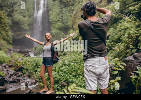 Eccitato giovane donna in piedi di fronte ad una cascata di essere fotografato con il suo fidanzato. Fotografo a scattare foto di donna. Foto Stock