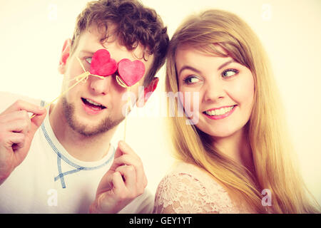 Amore e felicità concetto. Allegro piacevole giovane coppia con piccolo cuore su bastoni che copre donna uomo agli occhi. Gli amanti accecati dal loro grande amore. Foto Stock