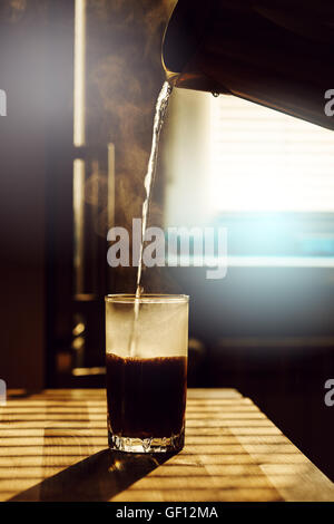 Acqua calda versare dal bollitore in vetro trasparente con la bevanda di caffè. Tavolo da cucina nella luce del sole. Immagine tonica, lente effetto flare Foto Stock