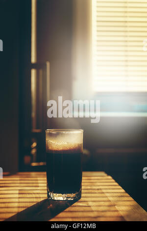 Vetro trasparente con caffè caldo drink sul tavolo da cucina nella luce del tramonto. Immagine tonica, lens flare effetto aggiunto. Foto Stock