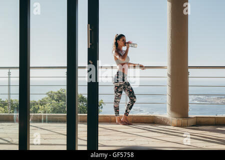 Foto all'aperto di una giovane donna in piedi sul balcone e beve acqua dalla bottiglia. donna in forma fisica che si prende una pausa dall'allenamento. Foto Stock