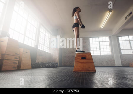 Bassa angolazione di montare i giovani modello femminile in sports wear in piedi su una scatola in palestra. Donna muscolare casella facendo saltare esercizio in palestra. Foto Stock