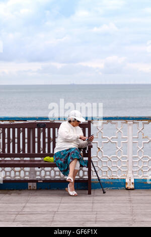 Donna anziana che interagisce con il suo dispositivo mobile sul Llandudno Pier, Wales, Regno Unito Foto Stock