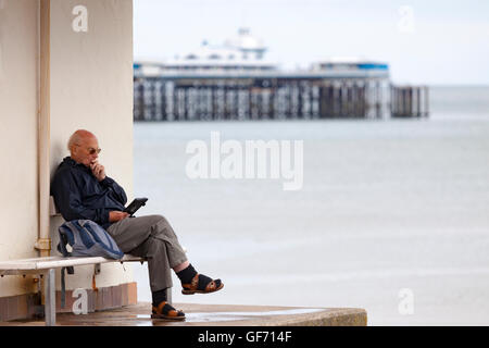 Uomo anziano si siede sotto un riparo promenade mentre la lettura del suo libro elettronico con il Molo di distanza, Llandudno, Galles Foto Stock