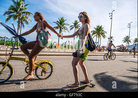 RIO DE JANEIRO - Marzo 6, 2016: giovane donna brasiliana su matasse di bicicletta un amico su skateboard sul fronte spiaggia di Ipanema road. Foto Stock