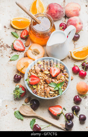 Sana colazione ingredienti. Oat granola nella ciotola con dadi, fragola e menta, il latte nel bricco, miele in un barattolo di vetro, frutta fresca, frutti di bosco in calcestruzzo leggero sfondo Foto Stock