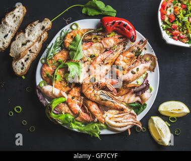 Piatto di gamberoni tigre arrostiti e pezzi di polipo con porri freschi, insalata, peperoni, limone, pane, pesto su sfondo nero Foto Stock